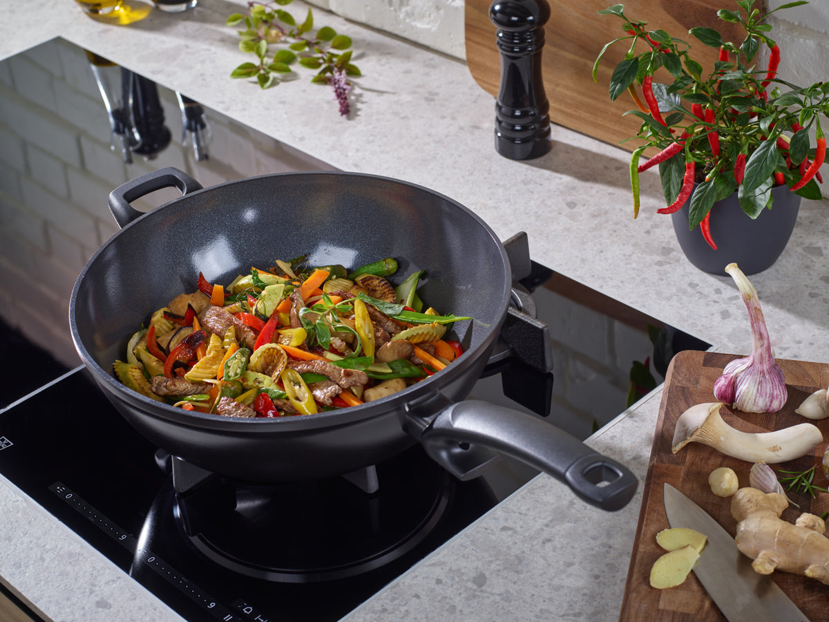 Ceratal® Wok Orbitschwarz von Fissler, PTFE-frei, auf dem Herd mit Rührbraten; frische Kräuter und Gewürze auf moderner Arbeitsplatte arrangiert.