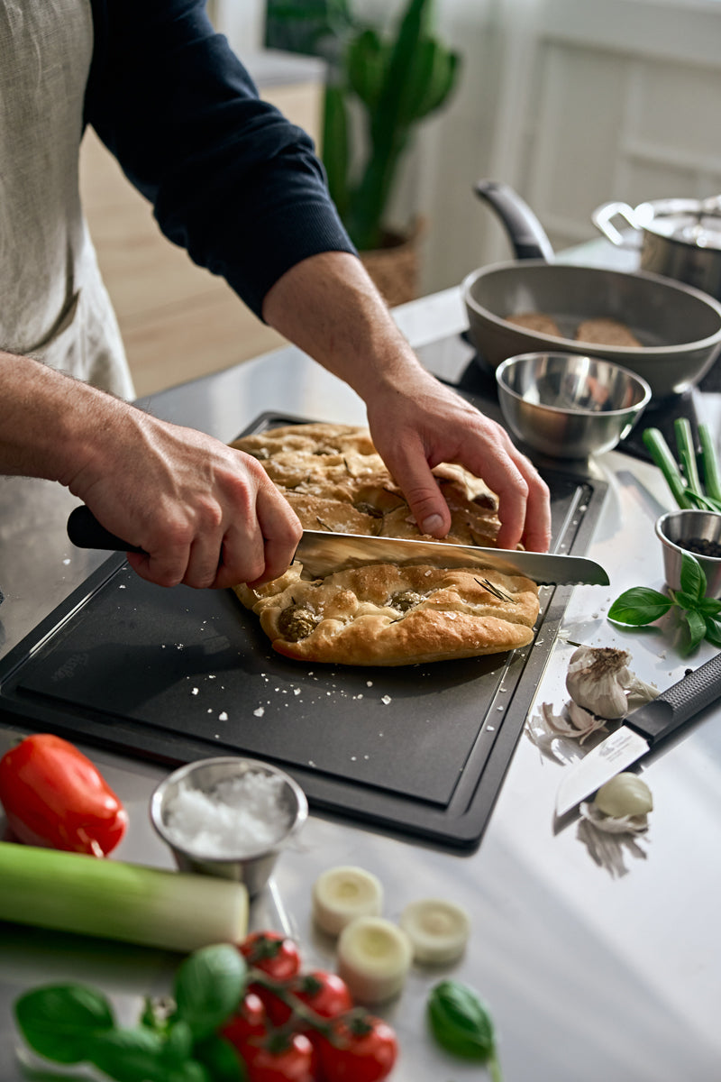 In einer sonnenbeschienenen Küche wird Focaccia mit dem Fissler ProCut Brotmesser 21cm geschnitten, inmitten von frischen Zutaten und modernem Design.