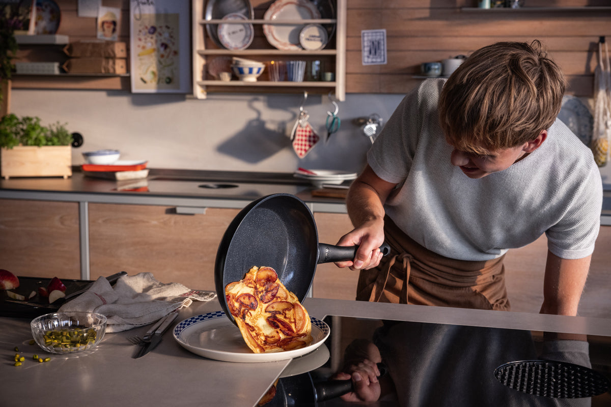 A person serves an omelet from a Fissler frying pan on a plate, surrounded by high-quality Fissler cookware and kitchen utensils.