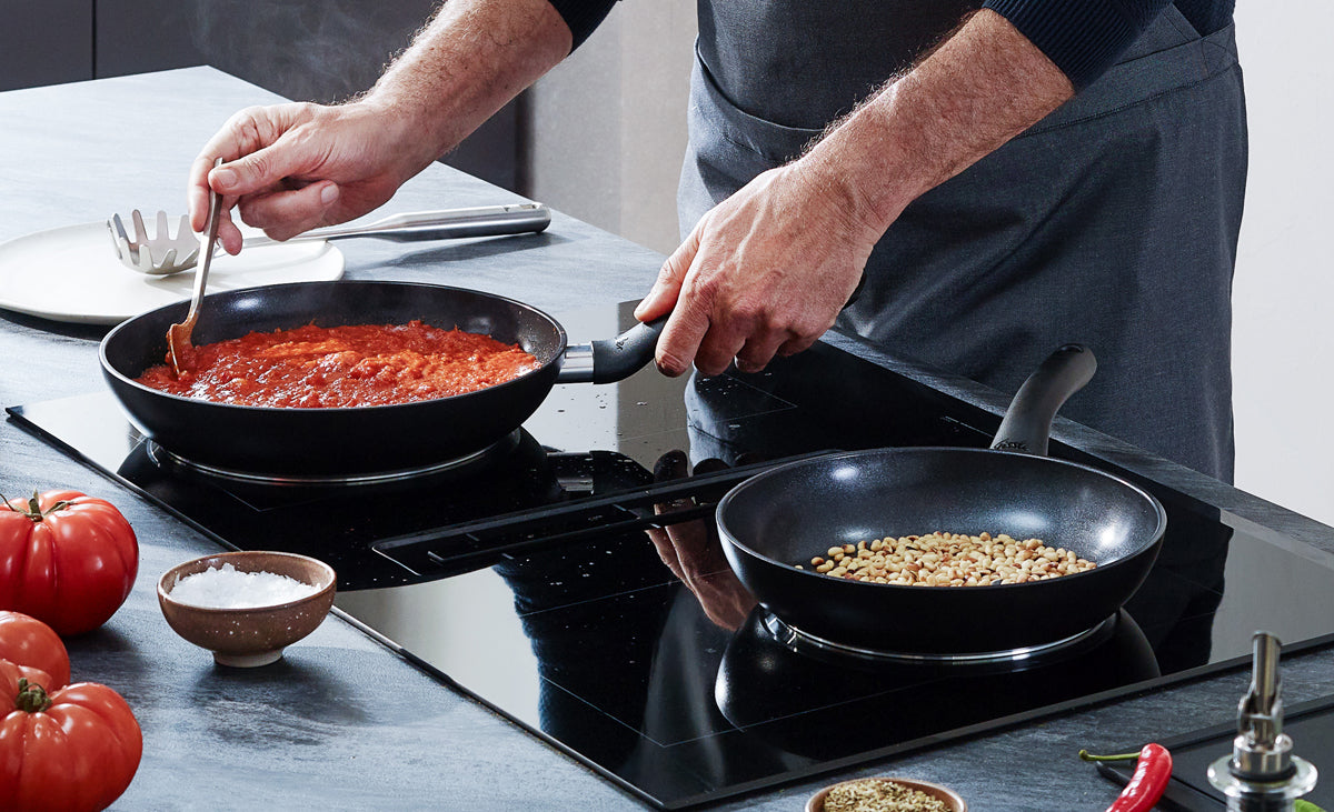 Ein Koch bereitet Tomatensauce zu und röstet Pinienkerne mit Fissler Essential® Pfannen und Küchenhelfer Edelstahl auf der Theke.