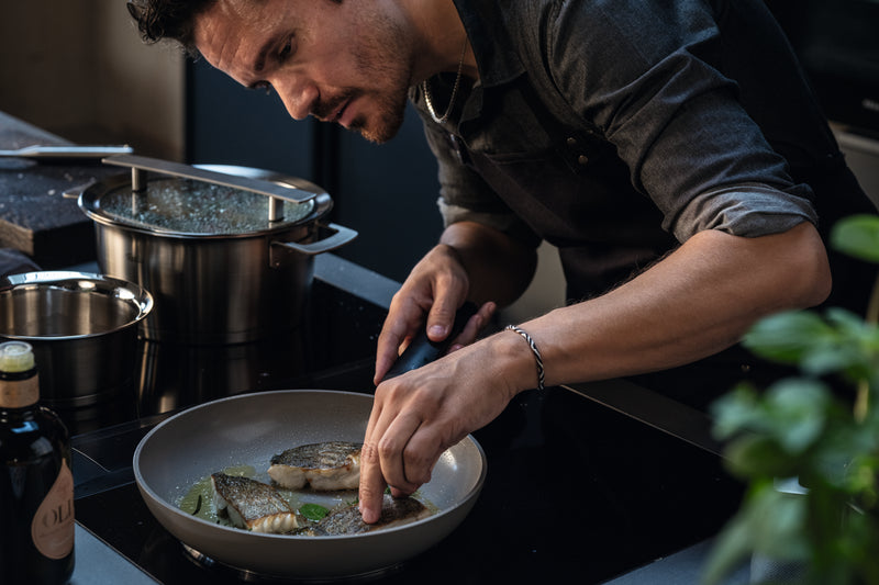 Ein Mann brät Fischfilets mit einer Zange in einer Fissler-Pfanne. Ein Fissler-Topf mit Glasdeckel und eine Ölflasche stehen auf dem Tresen.