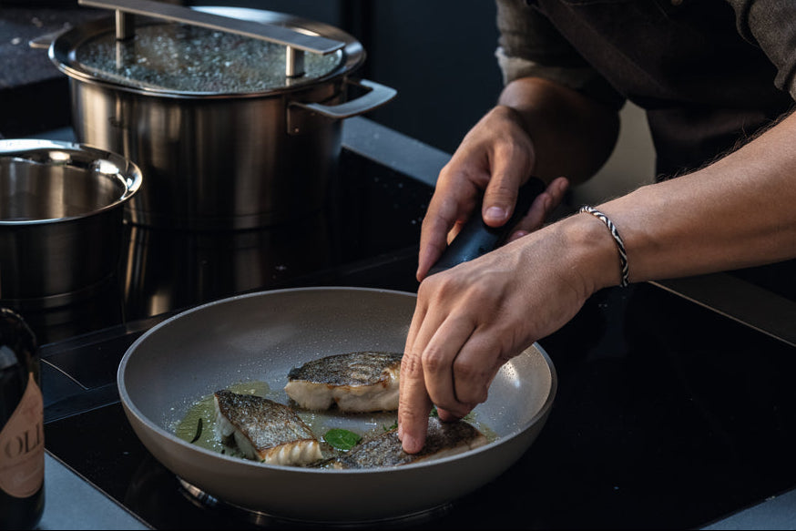 A person uses tongs to fry fish fillets in a Fissler frying pan; Fissler stainless steel pots sit on adjacent burners.