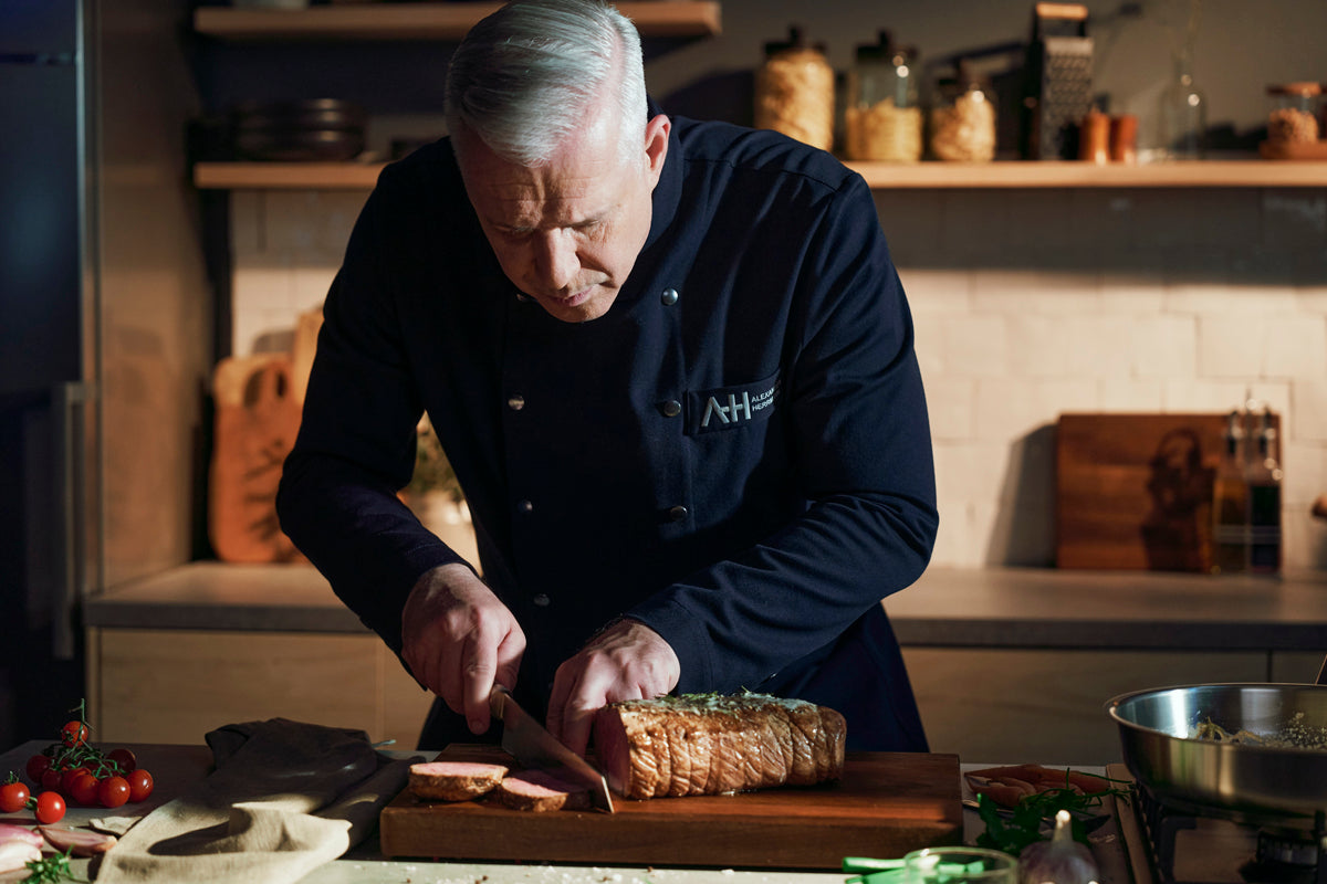 Ein Koch benutzt das Fissler PrimeCut 20cm Messer, um Brot auf Holz zu schneiden, umgeben von modernem Kochgeschirr in einer eleganten, organisierten Küche.