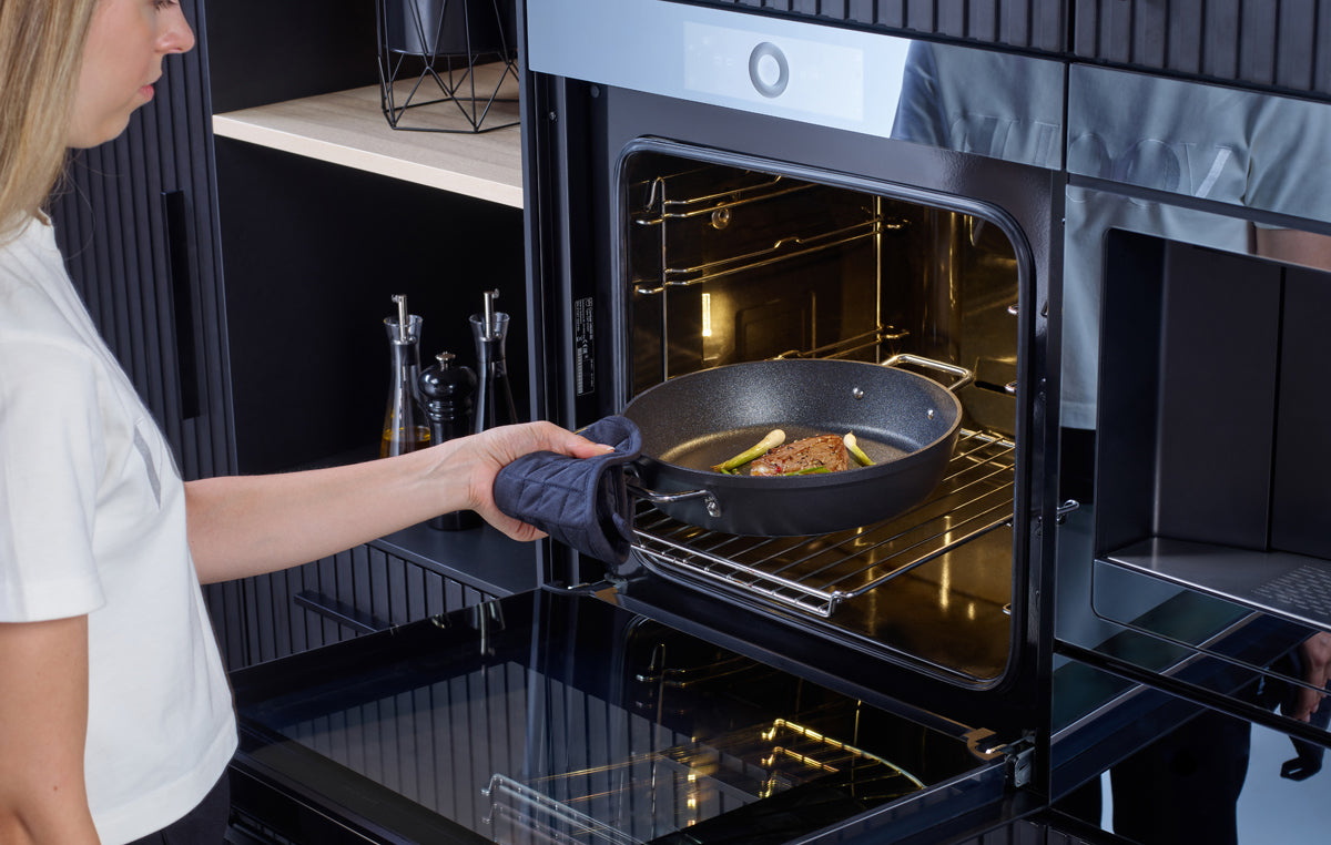 Eine Person benutzt einen Ofenhandschuh, um eine Fissler Adamant® Servierpfanne mit Fleisch und Gemüse in einen modernen Einbauofen zu stellen.