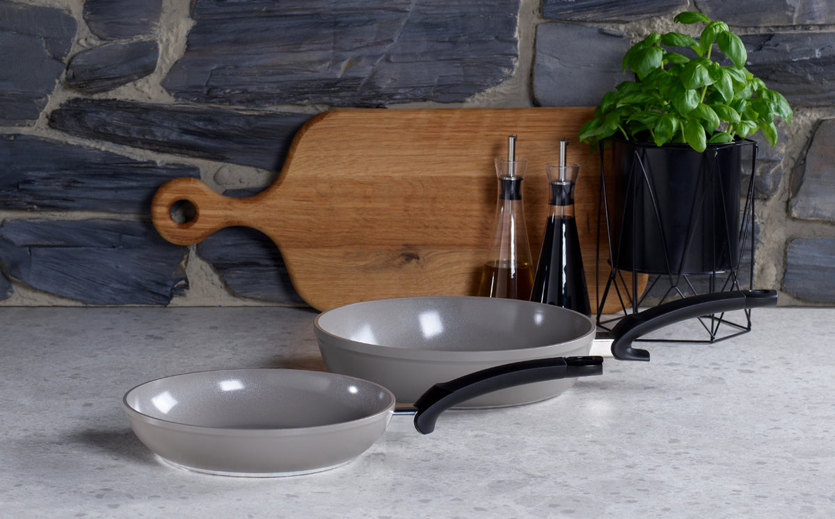Zwei Fissler Ceratal® Pfannen (28 cm & 24 cm) mit schwarzen Griffen auf einer Küchenzeile. PTFE-frei, Qualität Made in Germany.