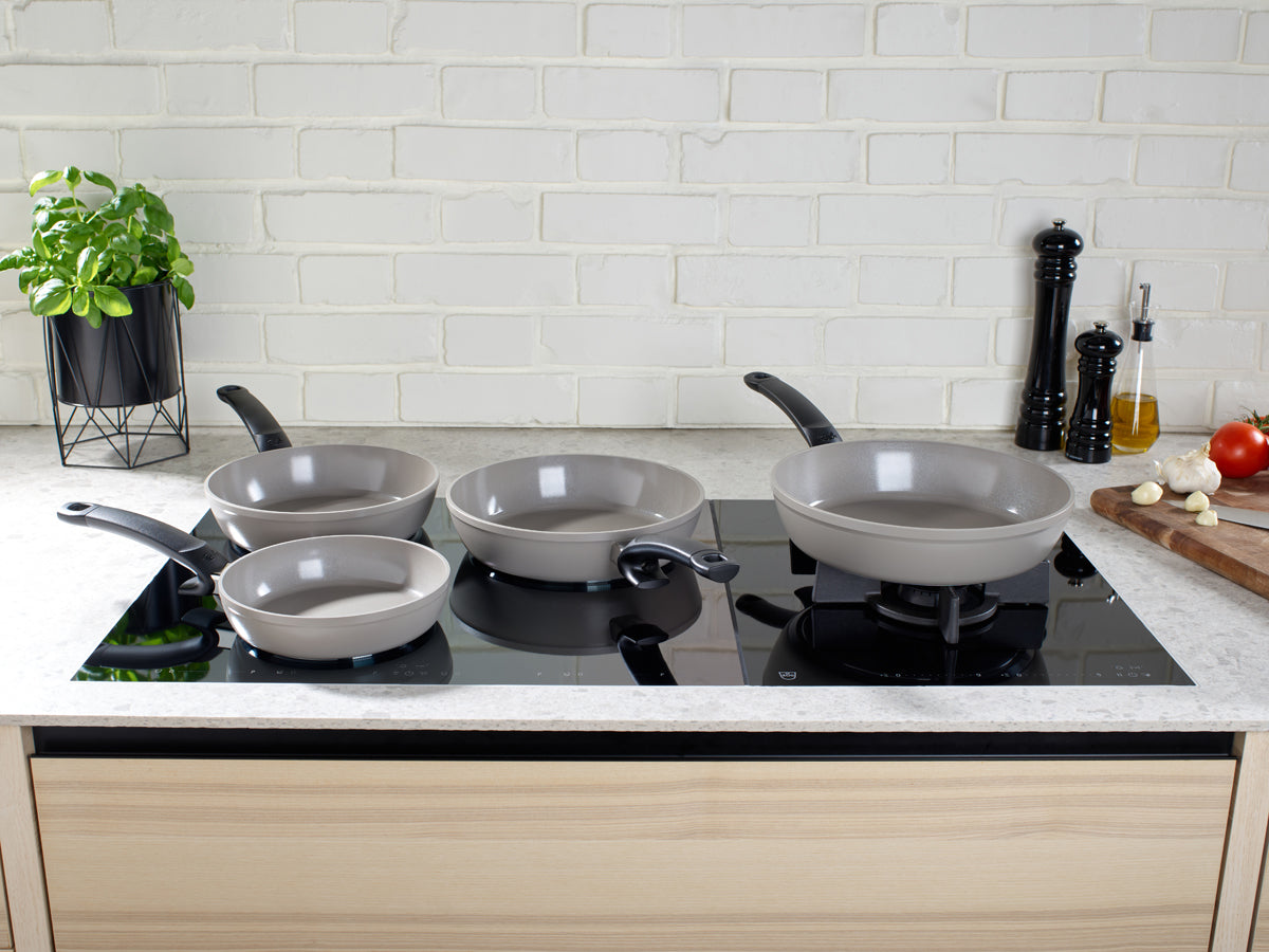 Two gray Fissler Ceratal Comfort pans (24cm & 28cm) with ceramic coating on a black induction hob, surrounded by fresh herbs.