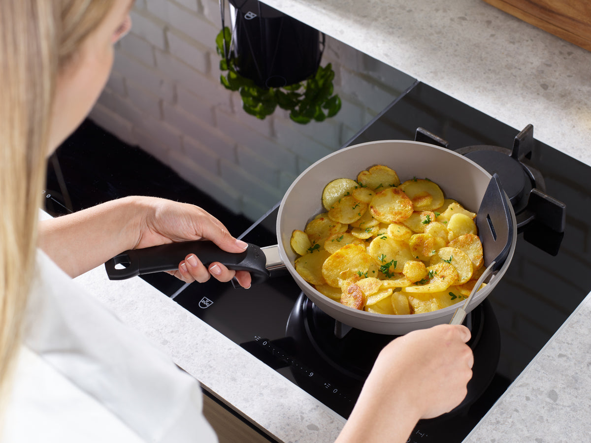 Goldbraune Kartoffeln brutzeln in einer Fissler Ceratal® Comfort Pan - PTFE-frei, made in Germany - auf einer glatten Herdplatte in einer hellen Küche.