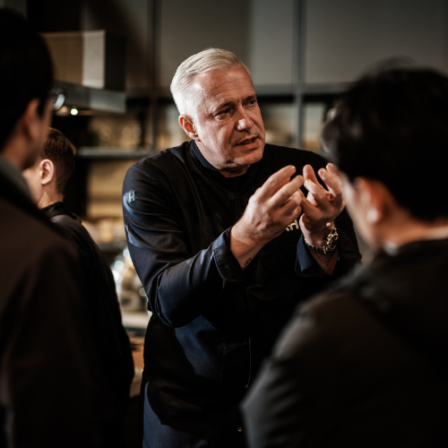 A chef with short gray hair passionately explains cookware techniques to a small group in a sleek, modern kitchen, gesturing with his hands as Fissler stainless steel pots and pans showcase German precision engineering on the countertop.