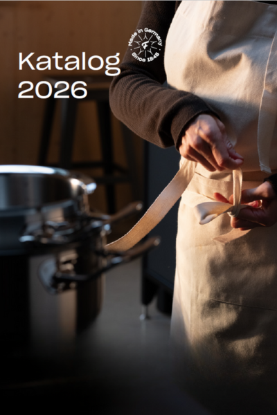 Eine Person in einer weißen Schürze bindet sich in der Nähe eines Fissler-Edelstahltopfes die Taille zu. In der oberen linken Ecke steht "Katalog 2026".