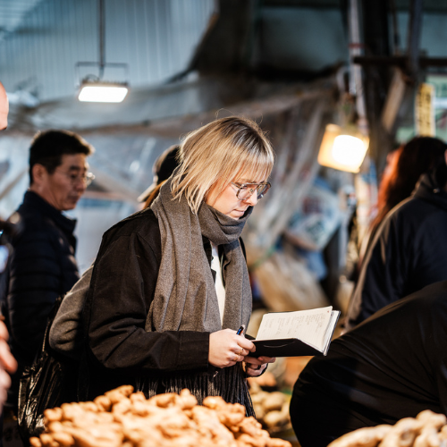 A person with blond hair and glasses writes in a notebook while standing at a market stall with an array of fresh mushrooms, evoking the inspiration behind exceptional cooking. Other shoppers and warm lights enhance the vibrant atmosphere, reflecting the attention to quality ingredients that pairs perfectly with Fissler's precision-engineered stainless steel cookware and timeless kitchen accessories.