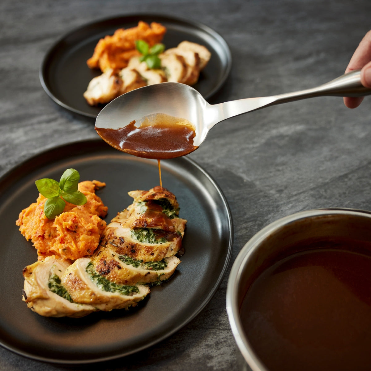 Eine Hand beträufelt mit dem Fissler Essential® Servierlöffel Gourmet-Hähnchen und Süßkartoffeln mit Sauce und zeigt damit deutsche Qualität.