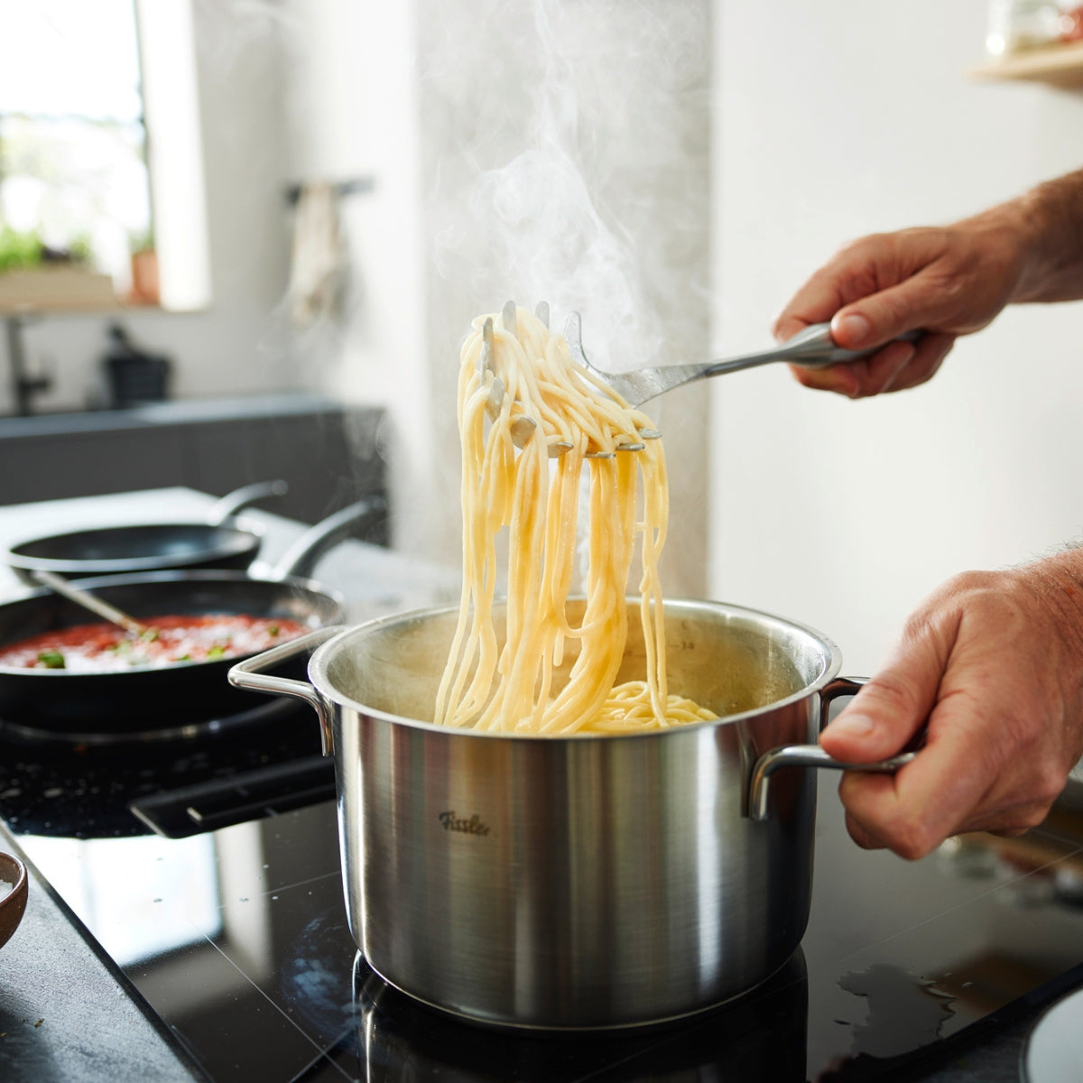 Gekochte Spaghetti werden mit dem Essential® Spaghettiheber aus einem dampfenden Fissler-Topf gehoben; Fissler-Pfanne auf dem Nachbarbrenner.