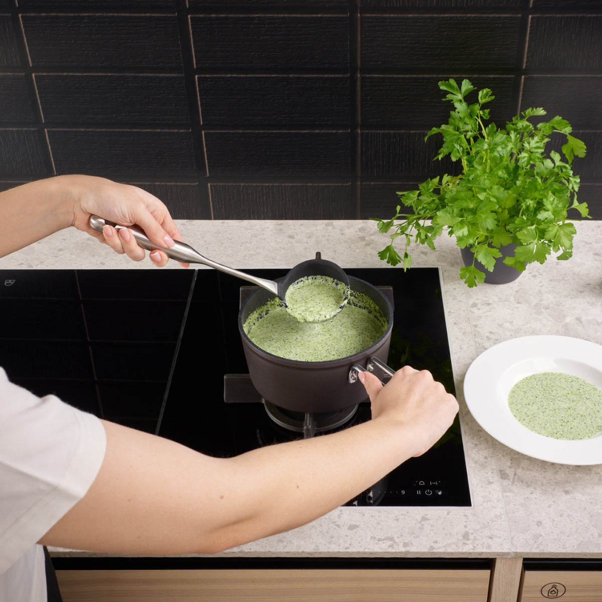 Eine Fissler Essential®-Silikonkelle serviert grüne Suppe aus einem Fissler-Topf in eine weiße Schüssel; daneben liegt frische Petersilie.