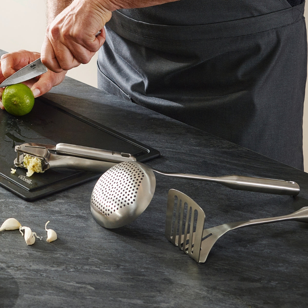 Ein Koch in einer grauen Schürze schneidet Limetten neben gehacktem Knoblauch, Nelken und Fissler Essential® Schaumlöffel auf einer glatten Arbeitsplatte.