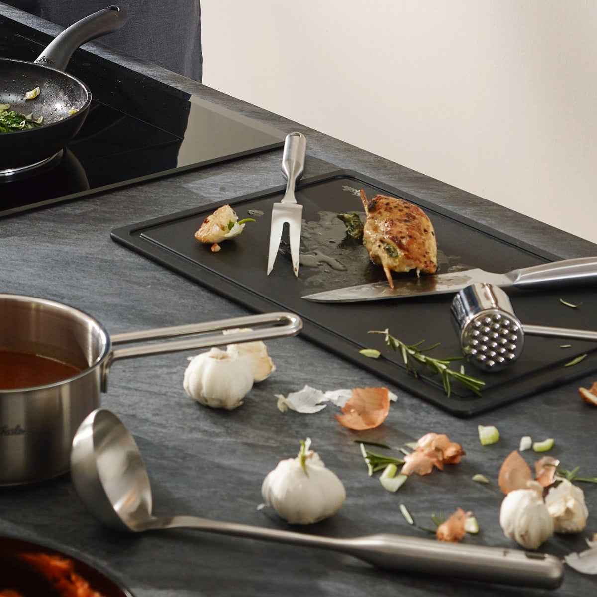 Gekochtes Hähnchen auf dem Brett mit Fissler Essential® Fleischgabel, Zartmacher; Fissler Kochtopf und Schöpfkelle auf dem Herd; moderne Küchenszene.