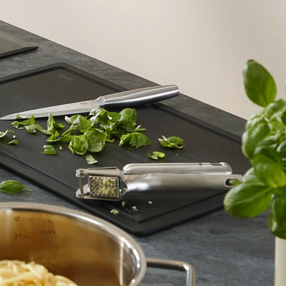 Fissler Essential® pot with pasta, garlic press, and fresh basil showcases German precision design on a modern kitchen counter.