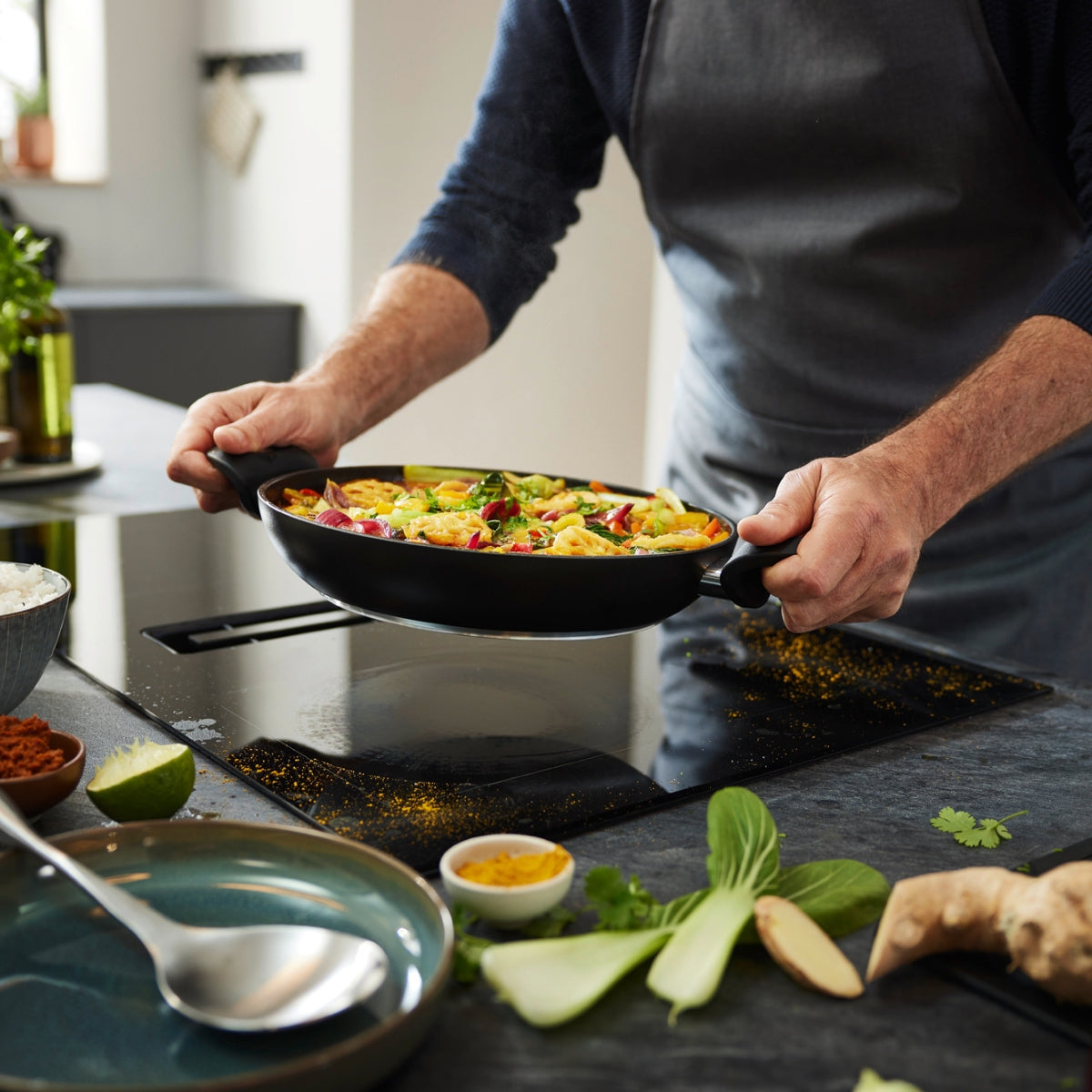 In einer eleganten Küche hält eine Köchin in grauer Schürze die Fissler Essential® Servierpfanne 28 cm und präsentiert ein farbenfrohes Wok-Gericht.
