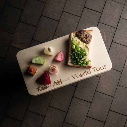 A wooden board labeled World Tour displays a carefully arranged slice of bread with fresh greens, assorted pickled vegetables, and a small square of sushi, all set on a refined dark tiled surface—an inviting showcase for culinary creations crafted with Fissler’s precision-engineered kitchenware.