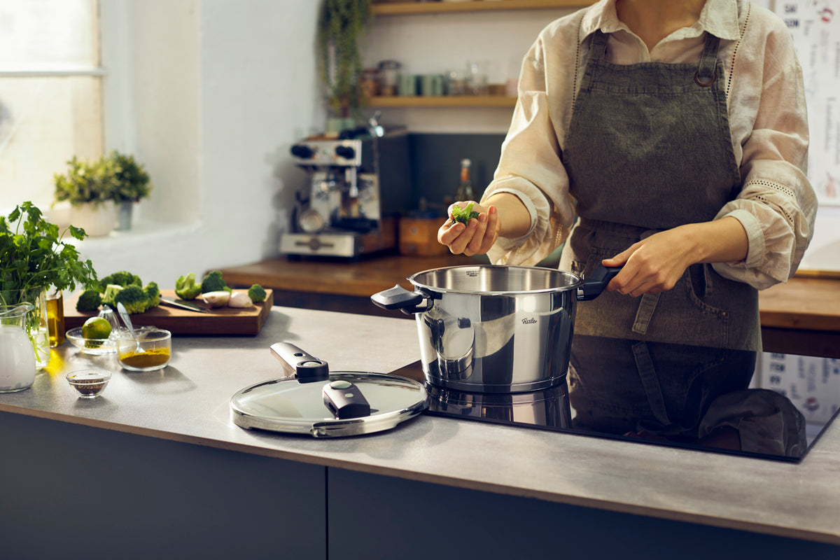 In einer Küche wird der Brokkoli über den Fissler Vitaquick® Elemental Schnellkochtopf gehalten, mit frischen Kräutern und Deckel auf der Theke.