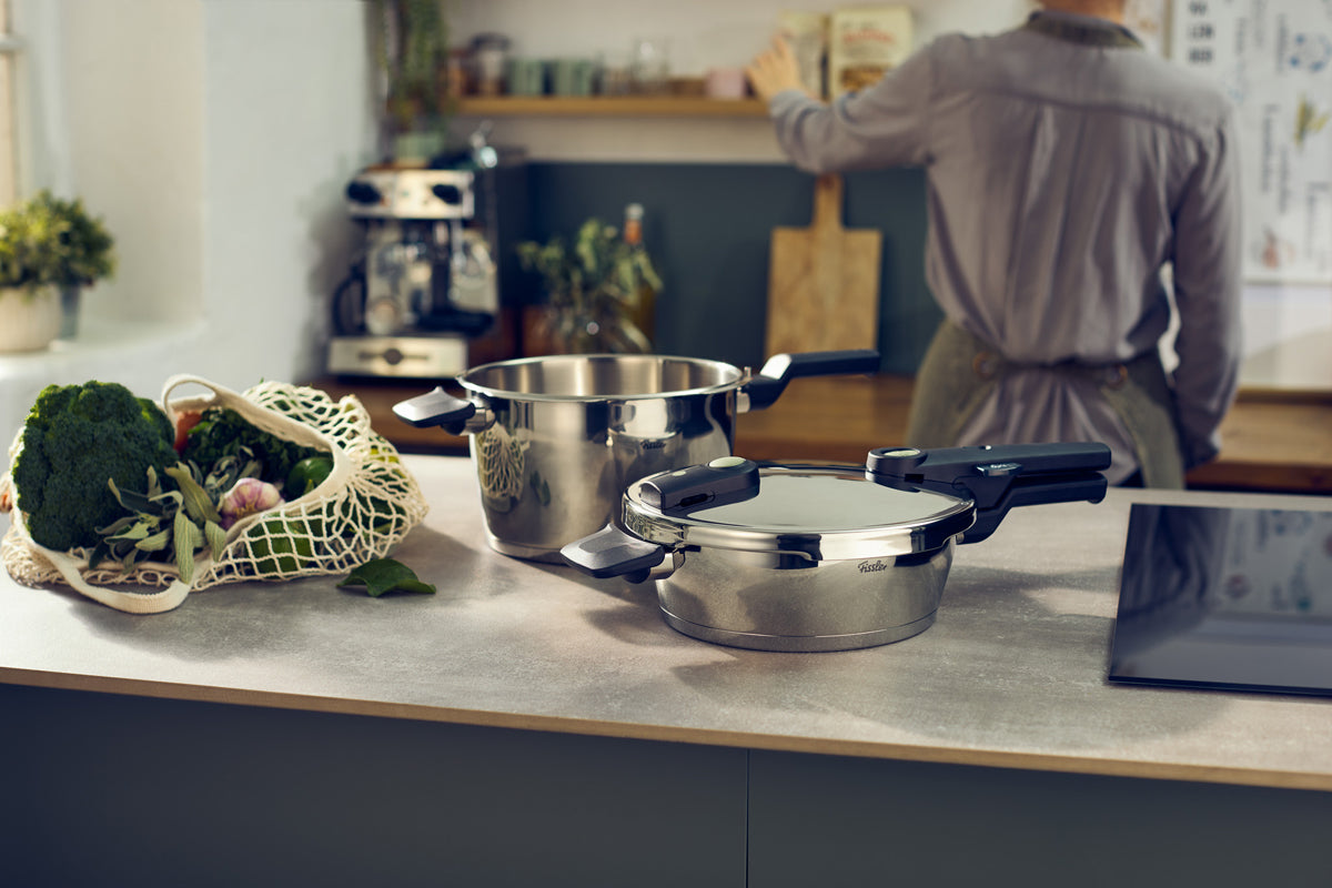 Fissler Vitaquick® Elemental-Töpfe (2,5 und 4,5 l) mit Deckeln auf einem Küchentisch, daneben Gemüse; Person im Hintergrund.