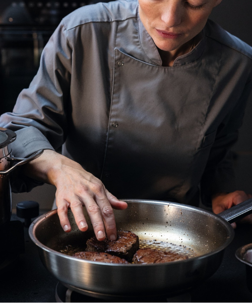 Ein Koch in grauer Uniform brät ein Steak in einer Fissler-Edelstahlpfanne an und zeigt damit deutsche Qualität und Innovation in einer Profiküche.