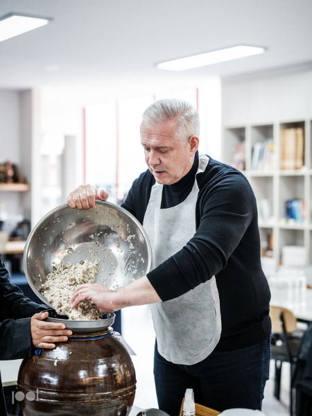In a bright, modern kitchen, a man in a crisp white apron pours a bowl of rice or grain mixture into a large ceramic jar. Sleek Fissler stainless steel cookware and premium German-engineered kitchen accessories can be seen on the counter, reflecting superior precision and timeless design.