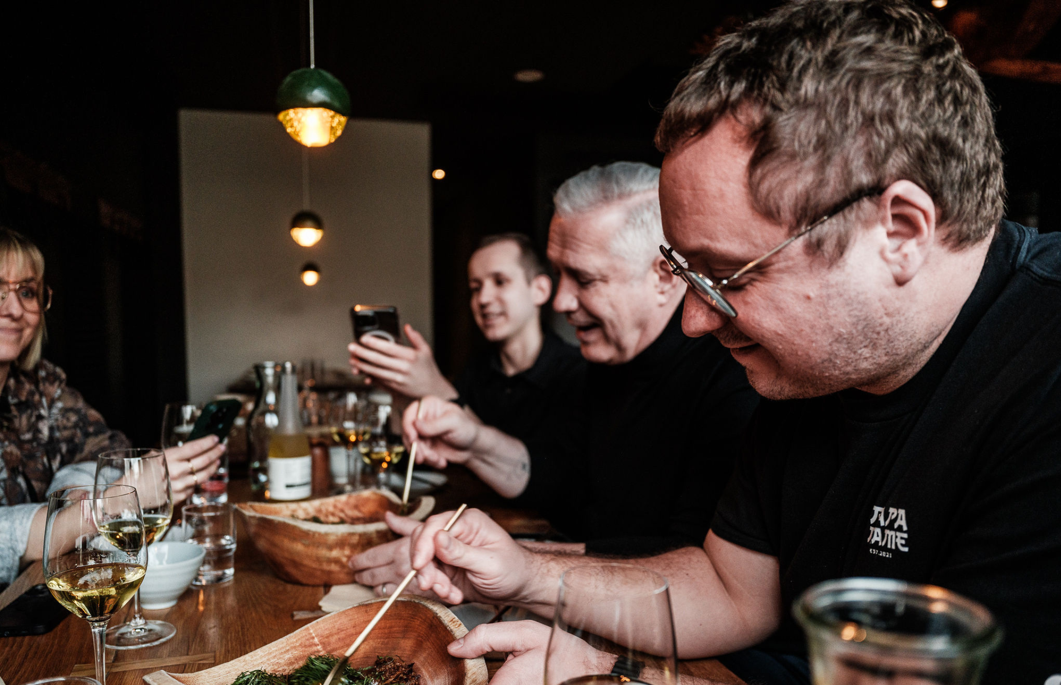Four people gather around a restaurant table, enjoying expertly prepared dishes with chopsticks. Two guests capture the moment on their phones, while Fissler’s elegant stainless steel cookware and serving accessories are featured among the wine glasses and plates on the wooden table—a reflection of precision engineering and timeless German design enhancing every dining experience.