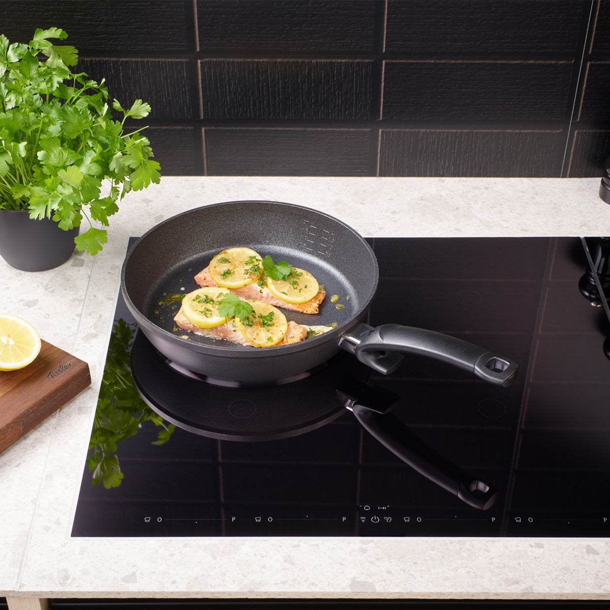 A premium Fissler frying pan, expertly engineered in Germany, sizzles with fish fillets and lemon slices atop a sleek black induction stove. Fresh herbs and a cut lemon rest on a wooden board beside the pan, all set against elegant dark tile—showcasing Fissler’s precision cookware in a timeless kitchen setting.