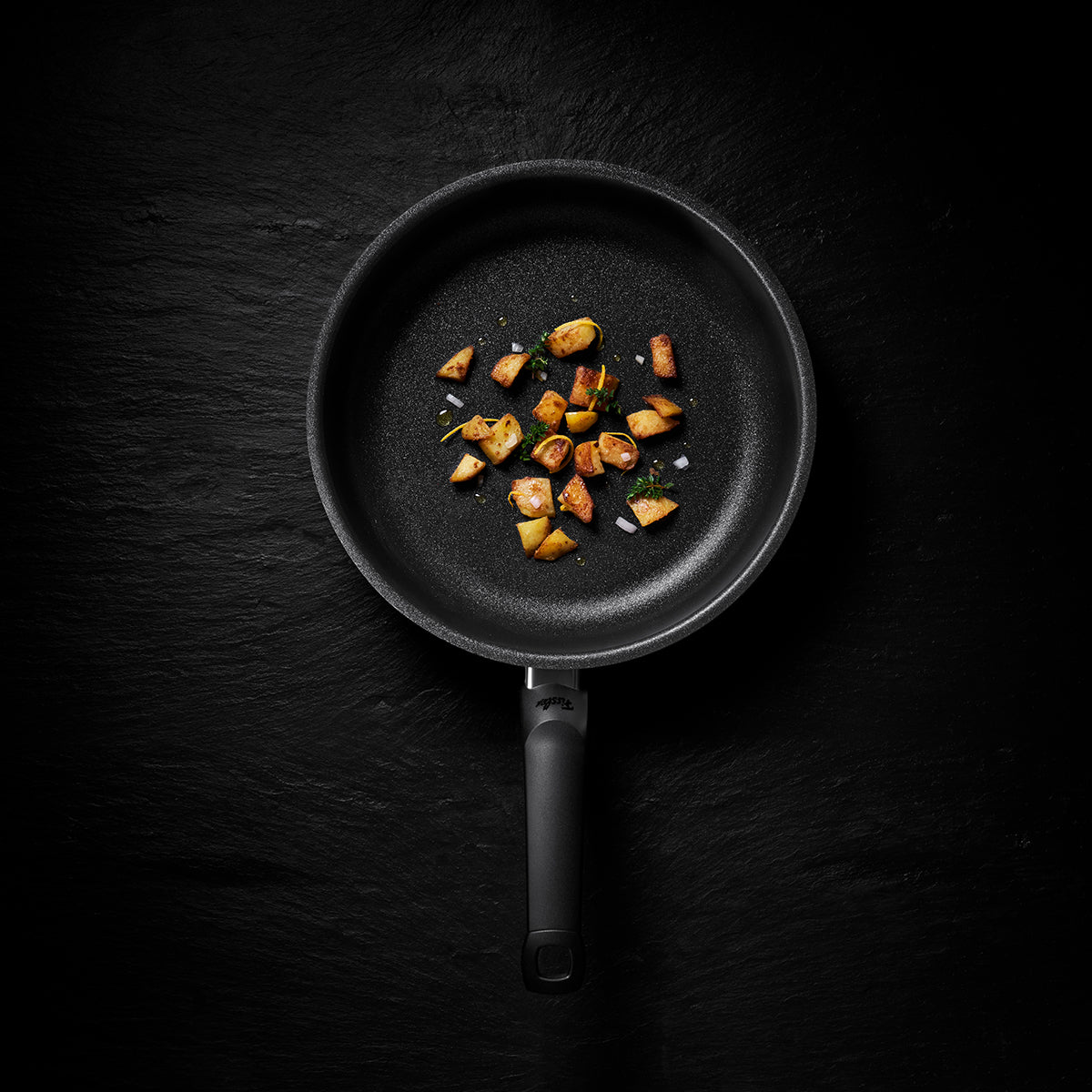A Fissler black frying pan, crafted with precision engineering for durability and timeless design, holds a few golden-browned potato cubes garnished with fresh herbs on a refined dark, textured surface—showcasing German quality and innovation in cookware.