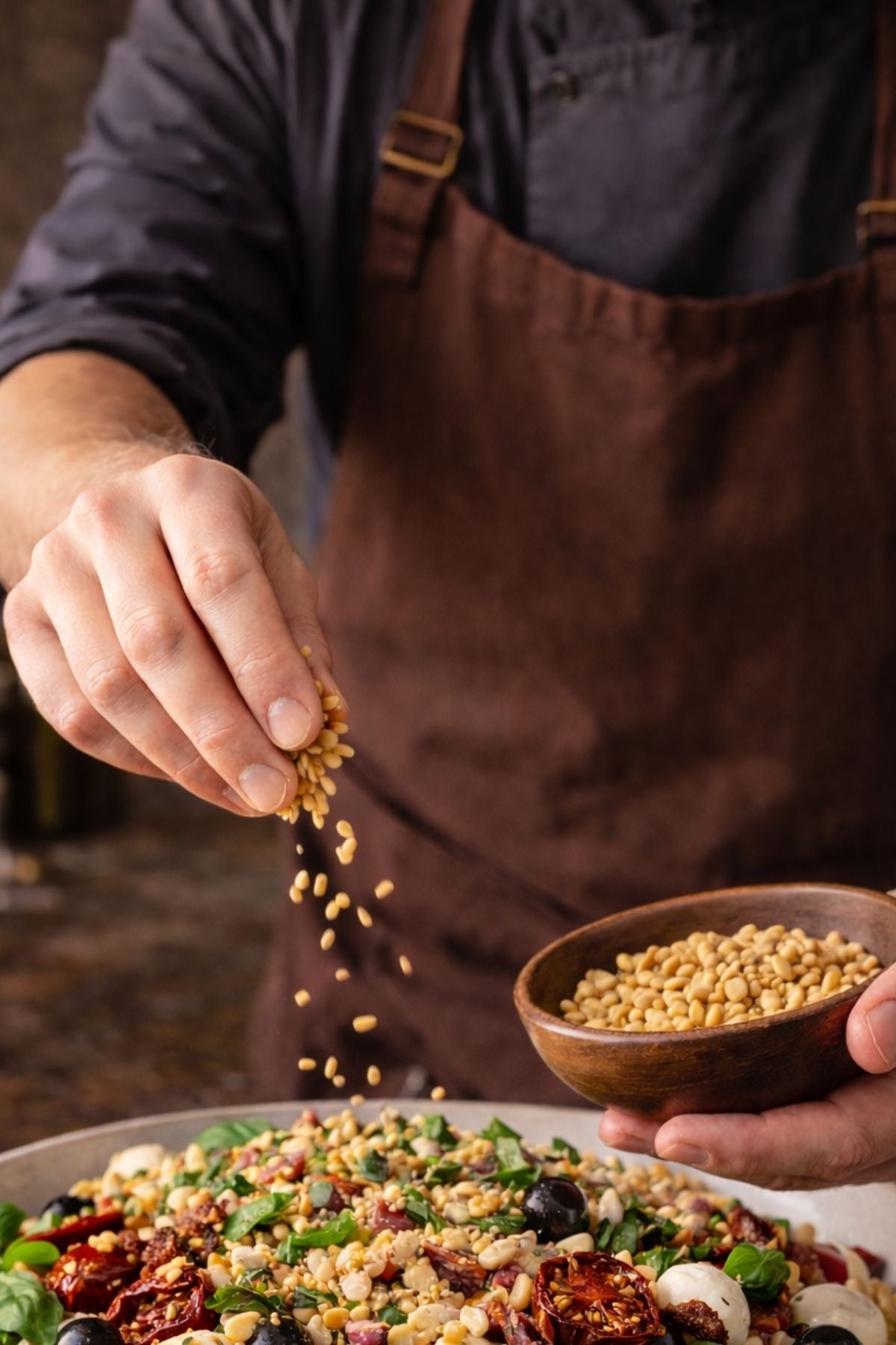 A chef in a brown apron adds pine nuts to a vibrant salad, using durable Fissler cookware, renowned for precision and timeless quality.