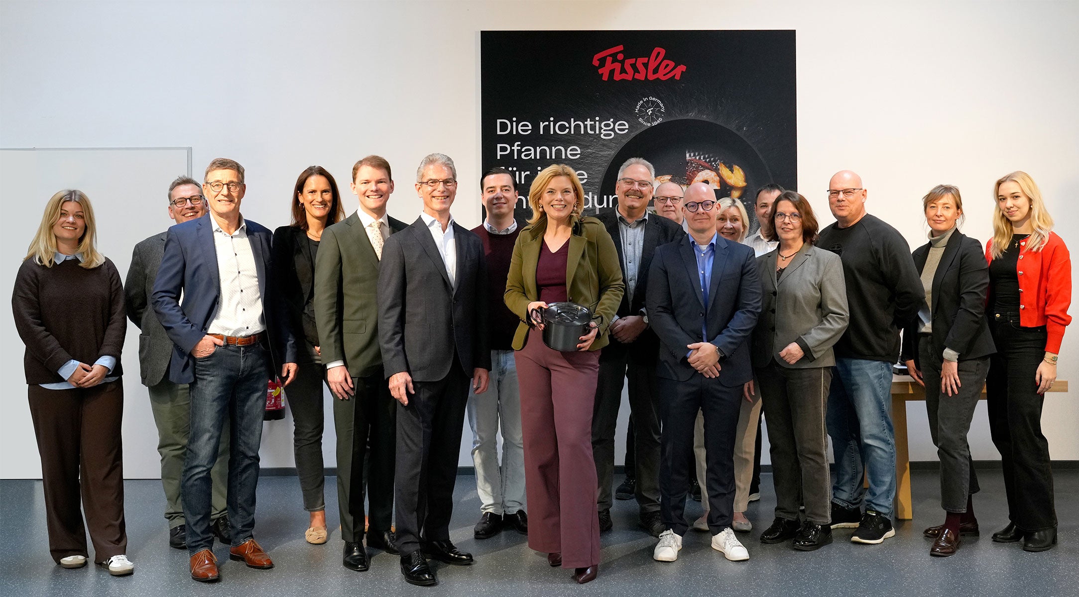 Fünfzehn Kollegen in Business Casual posieren neben dem Fissler-Logo; die Frau in der Mitte hebt einen hochwertigen Fissler-Kochtopf aus deutscher Produktion hervor.