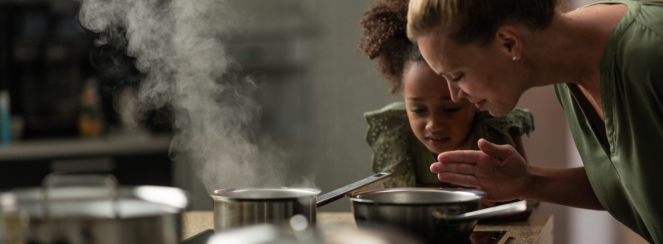 Eine Frau und ein junges Mädchen genießen das Aroma eines Fissler-Topfes, umgeben von elegantem Fissler-Kochgeschirr aus Edelstahl in einer modernen Küche.