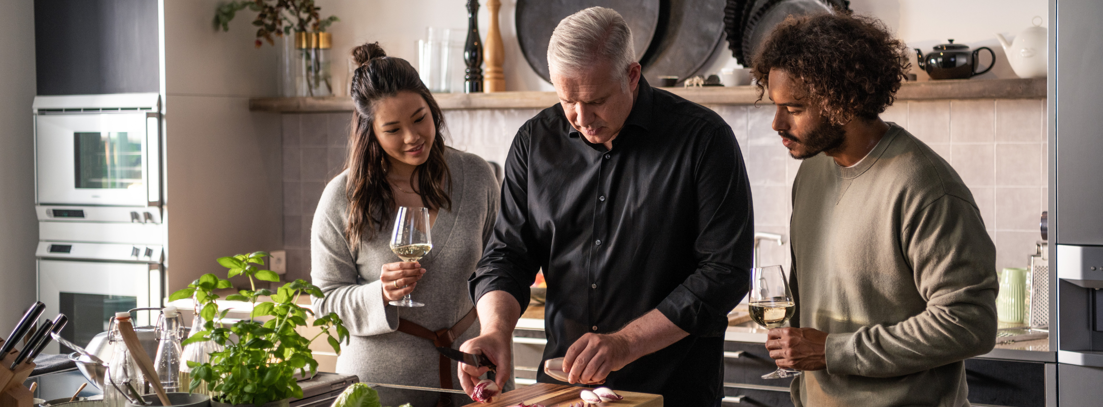 Ein Mann schneidet Gemüse mit Fissler-Kochgeschirr, während sich Freunde in der Nähe unterhalten, umgeben von eleganten Küchenutensilien aus deutscher Produktion.
