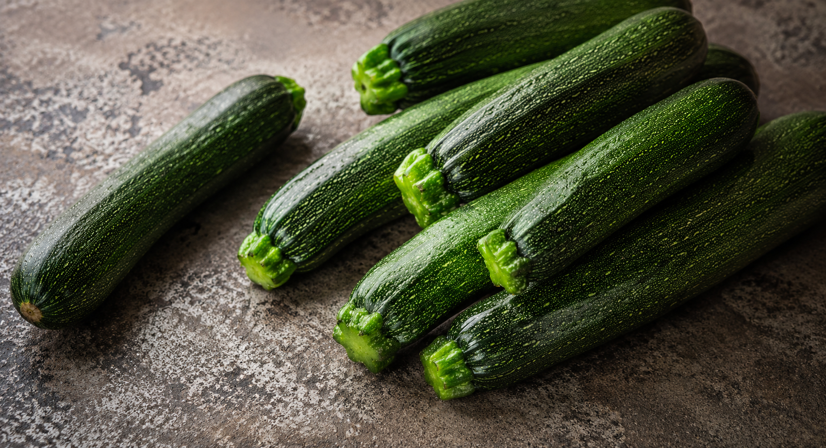 A selection of vibrant green zucchinis rests on a speckled brown countertop, highlighting the ideal fresh ingredients to prepare in Fissler’s precision-engineered stainless steel cookware. One zucchini lies slightly apart, exemplifying the attention to detail and quality that defines our German-made kitchen accessories and cooking tools since 1845.