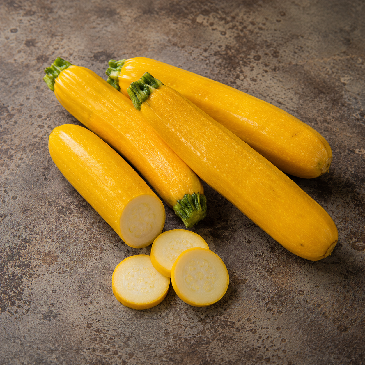Four whole yellow zucchinis rest on a stone surface, with one precisely sliced to display its pale interior. In front, three neatly cut zucchini rounds are arranged, embodying the freshness and quality ingredients perfect for preparing with Fissler’s expertly engineered cookware.