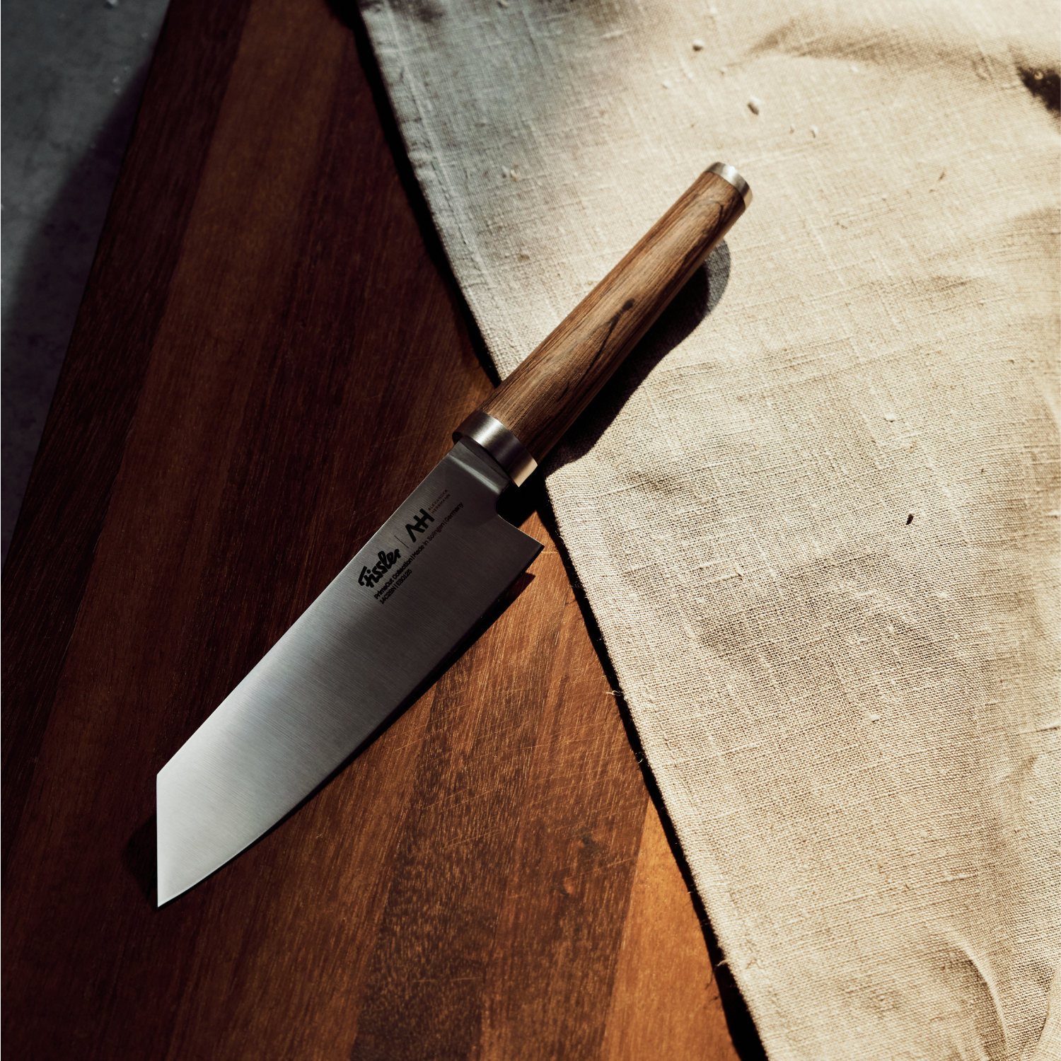 A Fissler chef's knife with a handmade wooden handle on wood next to a beige linen, showcasing precise German design in dramatic light.