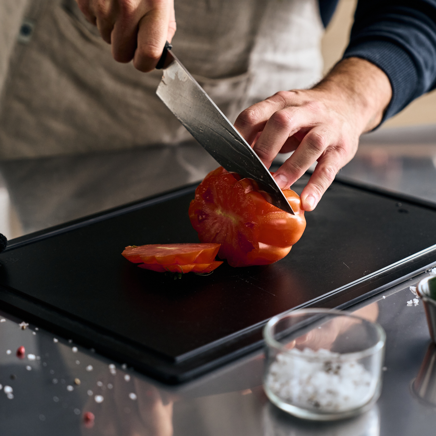 A person in an apron cuts a tomato on a high-quality Fissler cutting board. Flaky salt and a glass bowl are next to it.