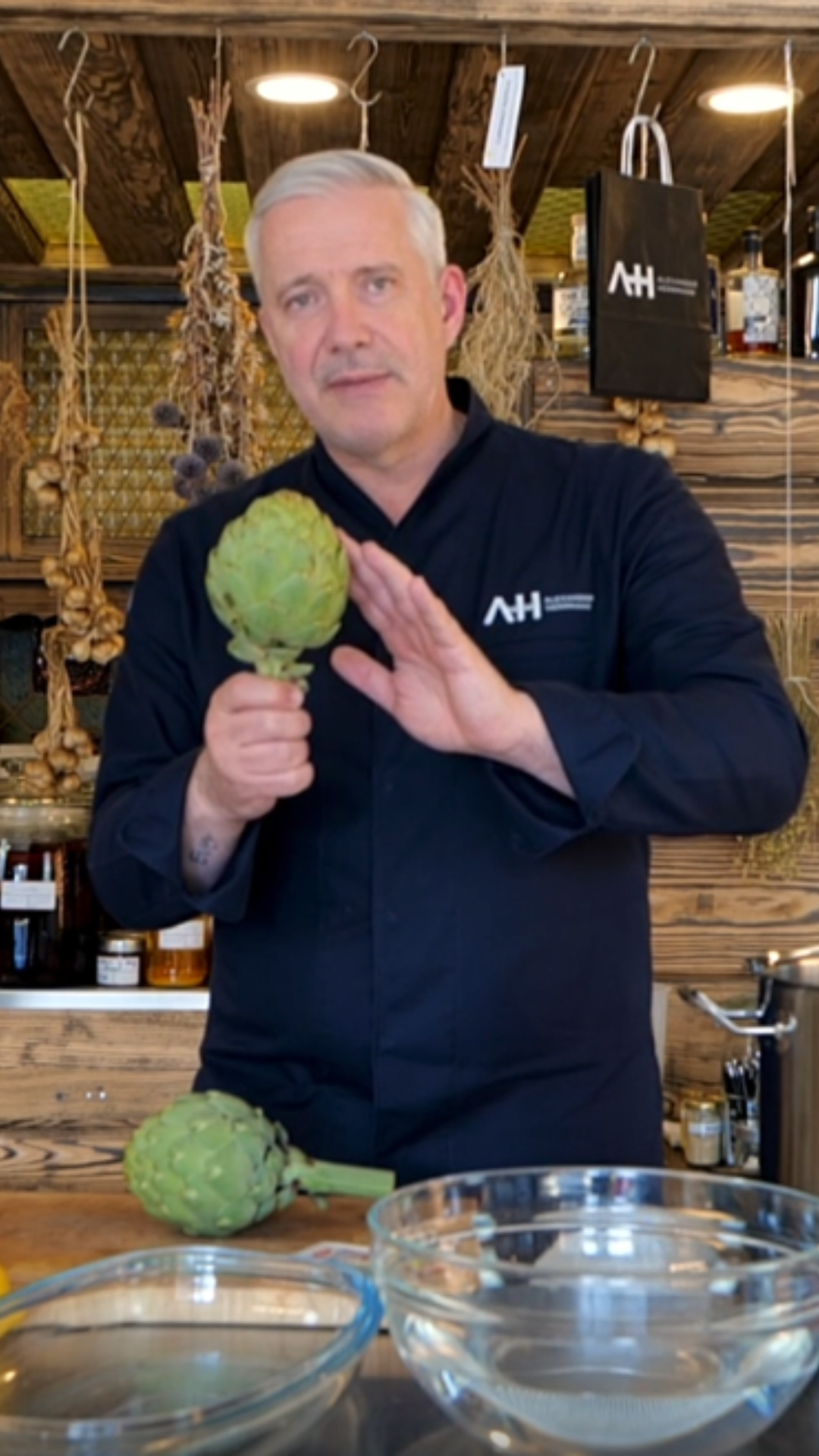 A chef in a rustic kitchen holds a fresh artichoke next to Fissler cookware and glass bowls; timeless tools hang in the background.