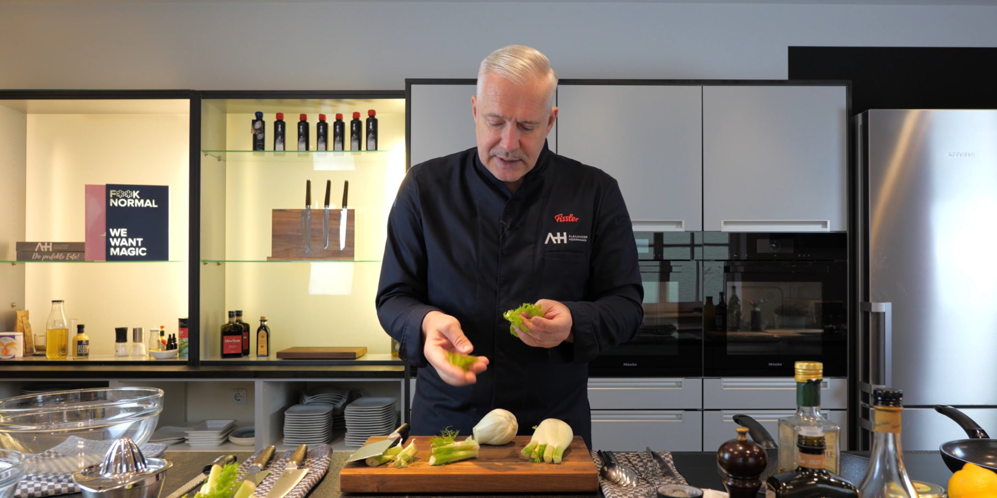 Ein Koch bereitet Fenchel auf einem Holzbrett in einer modernen Küche zu. Hinter ihm stehen hochwertige Fissler-Kochgeschirre und -Geräte.