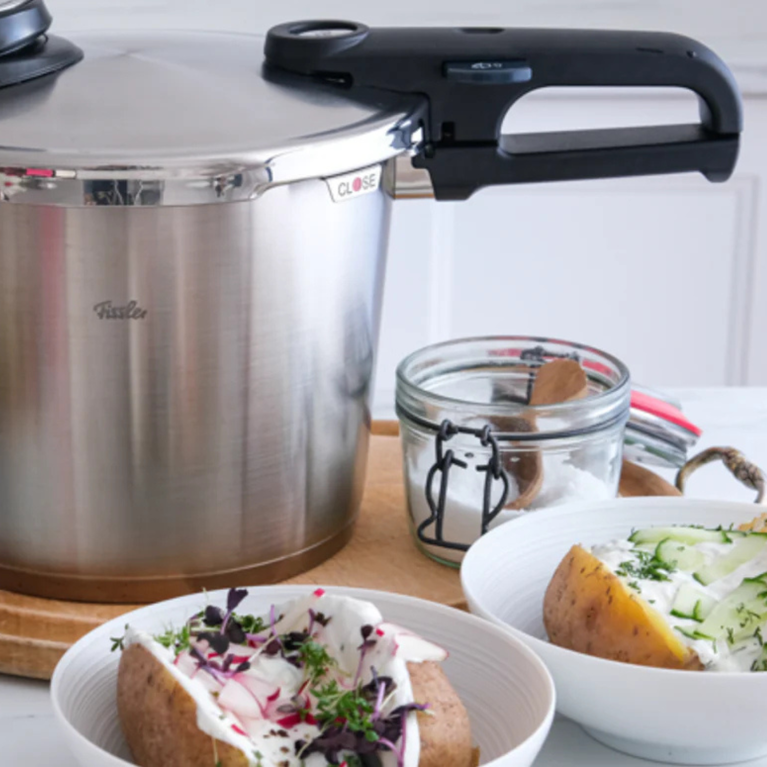 Ein Schnellkochtopf aus rostfreiem Stahl von Fissler auf einem Holzbrett, dazu zwei Schalen mit gebackenen Kartoffeln, die mit Kräutern und saurer Sahne belegt sind.