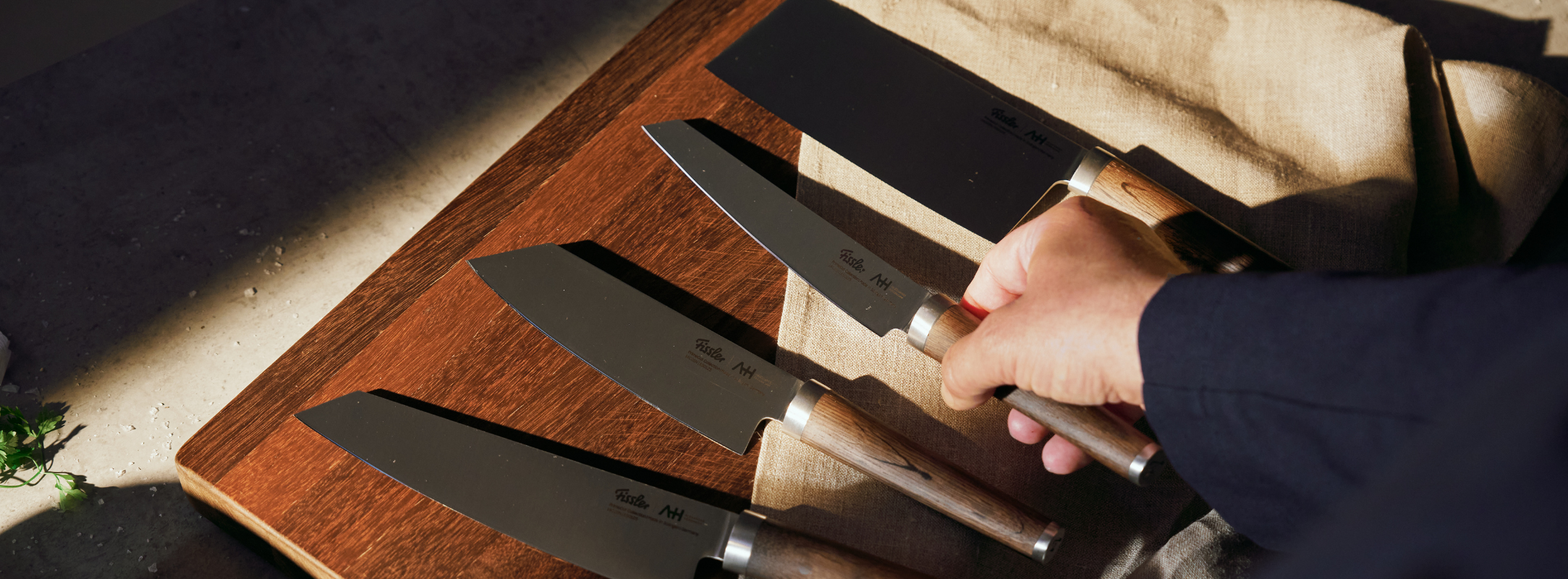 Eine Hand wählt ein Fissler-Kochmesser aus vier präzisionsgefertigten Messern mit Holzgriffen aus, die auf einer Tafel in warmem Tageslicht ausgestellt sind.