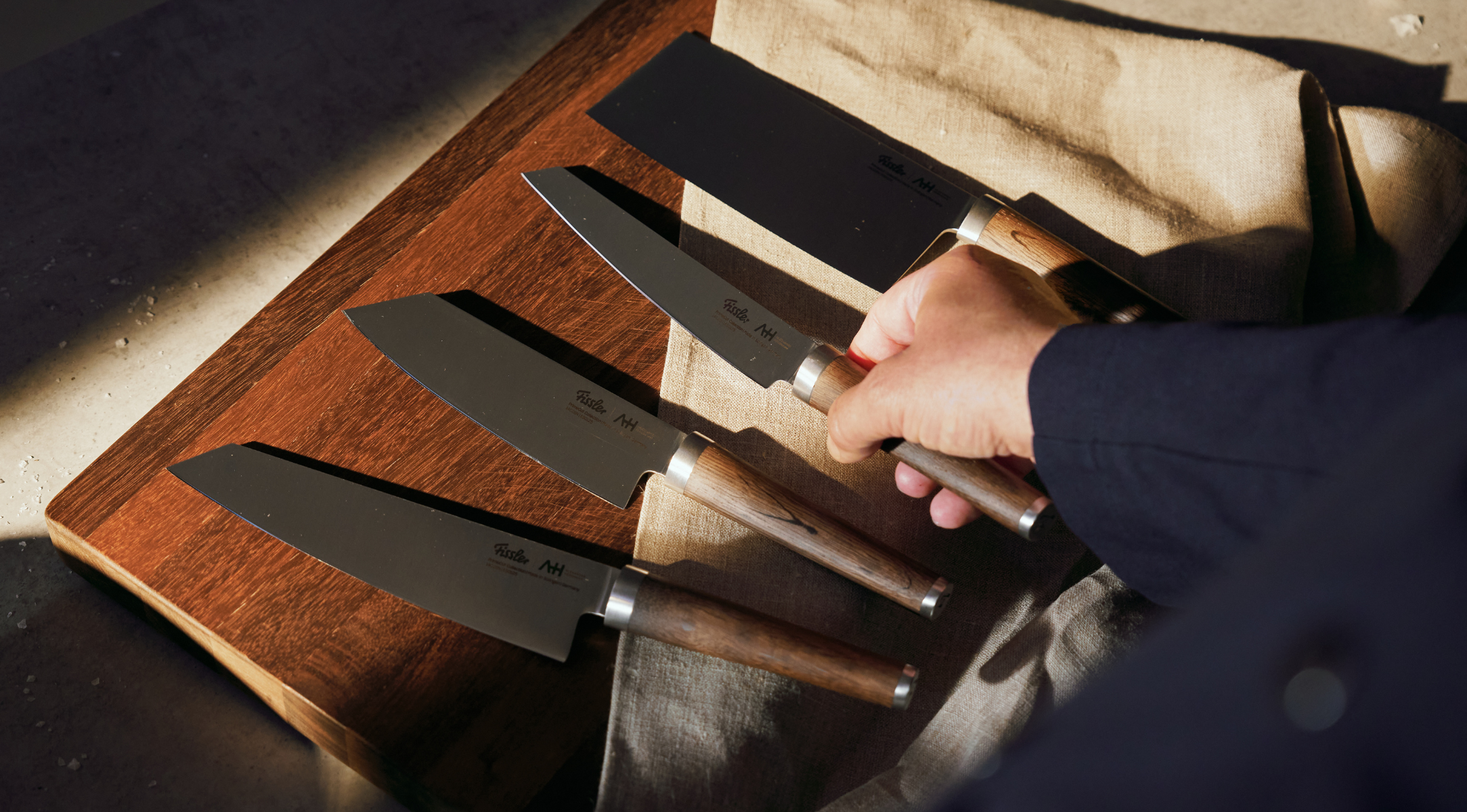 Eine Hand wählt ein Fissler-Küchenmesser aus vier hochwertigen Messern mit Holzgriffen auf einem Brett aus, das in natürliches Sonnenlicht getaucht ist.