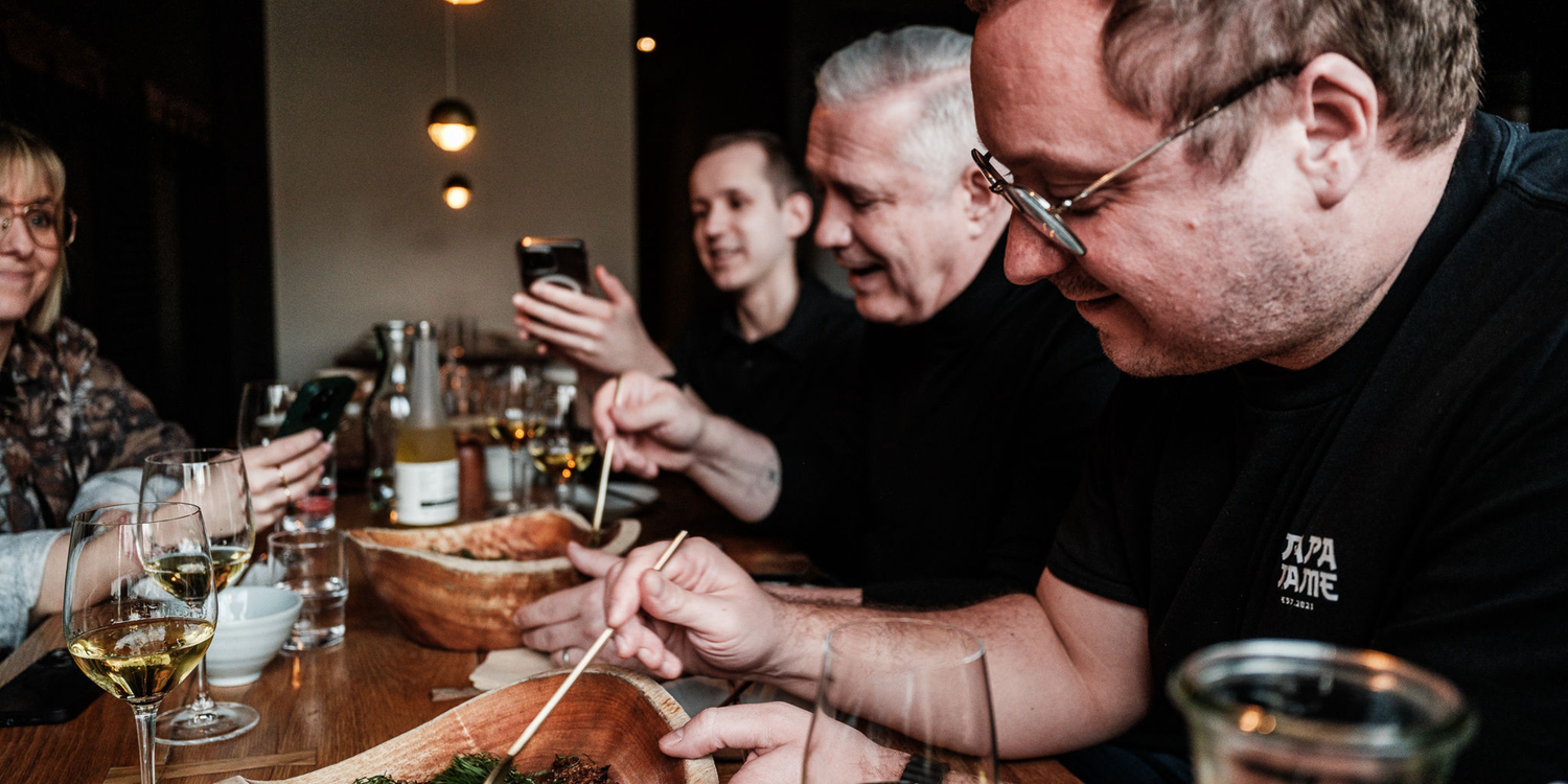 Vier Personen sitzen um einen hölzernen Restauranttisch herum, der mit köstlichen Speisen, Getränken und glänzendem Edelstahlgeschirr gedeckt ist, das das zeitlose deutsche Design von Fissler zur Geltung bringt. Die lächelnden Gäste essen mit Essstäbchen, während zwei von ihnen den Moment mit ihren Handys festhalten. Weingläser und -flaschen tragen zur einladenden, warmen Atmosphäre bei – sie stehen für Genuss, Langlebigkeit und innovative Präzision bei jeder Mahlzeit.