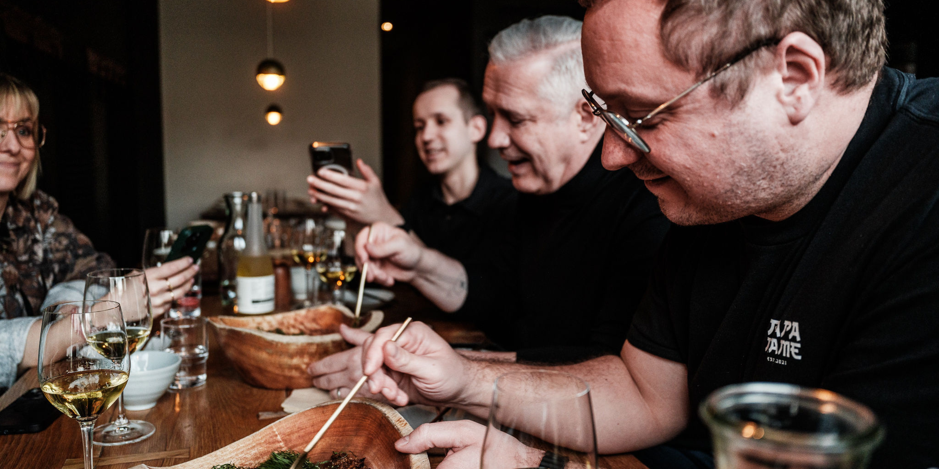 Vier Personen sitzen um einen hölzernen Restauranttisch herum, der mit köstlichen Speisen, Getränken und glänzendem Edelstahlgeschirr gedeckt ist, das das zeitlose deutsche Design von Fissler zur Geltung bringt. Die lächelnden Gäste essen mit Essstäbchen, während zwei von ihnen den Moment mit ihren Handys festhalten. Weingläser und -flaschen tragen zur einladenden, warmen Atmosphäre bei – sie stehen für Genuss, Langlebigkeit und innovative Präzision bei jeder Mahlzeit.