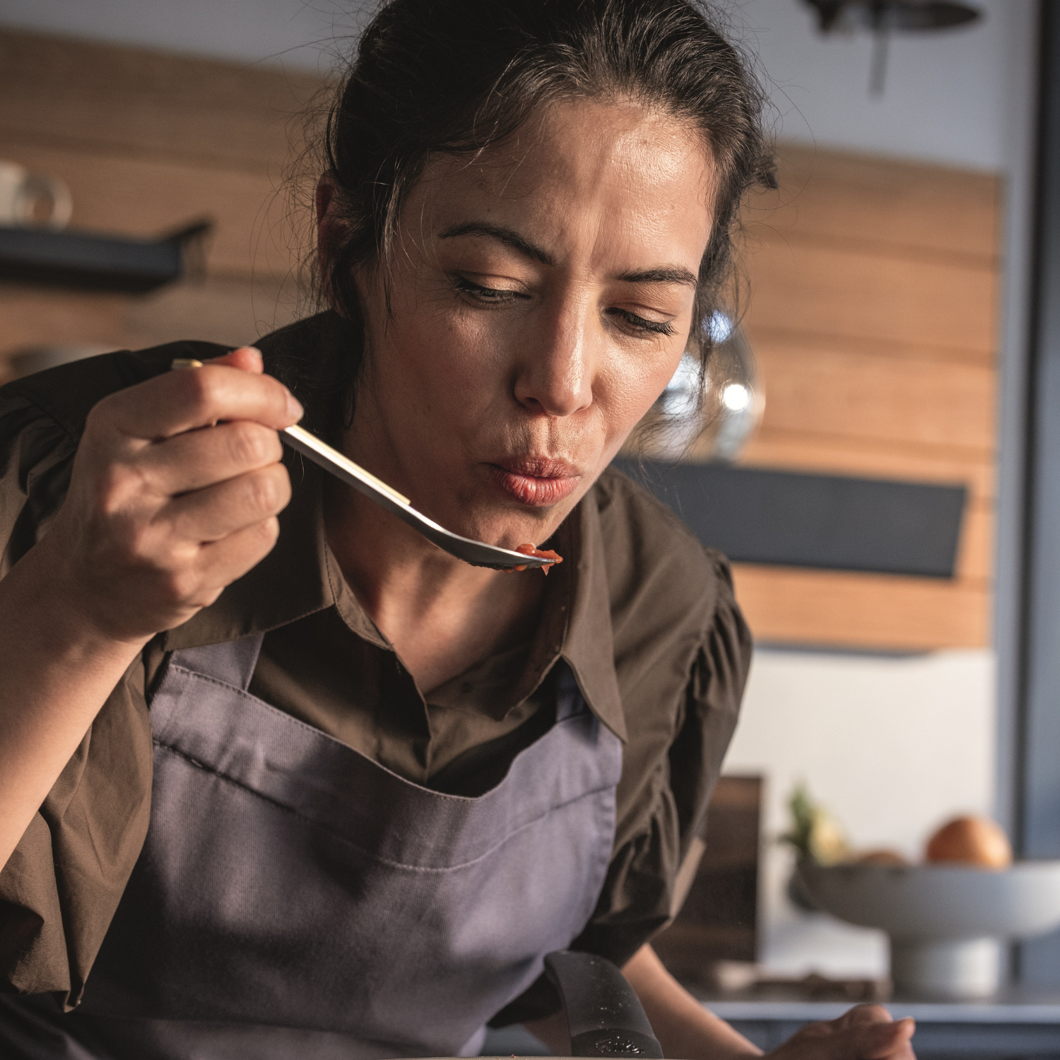 A woman in an apron tastes food from a spoon in a sleek, modern kitchen featuring premium Fissler stainless steel cookware. She appears focused and thoughtful as she evaluates the flavor, surrounded by expertly engineered pots and pans that showcase Fissler’s renowned German craftsmanship and timeless design.