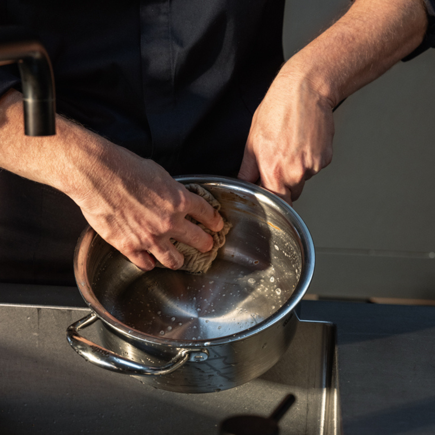 A person cleans a Fissler stainless steel pot with a sponge at the kitchen sink, demonstrating the ease of caring for our precisely engineered, German-made cookware. The sleek, durable design stands out even during everyday maintenance.