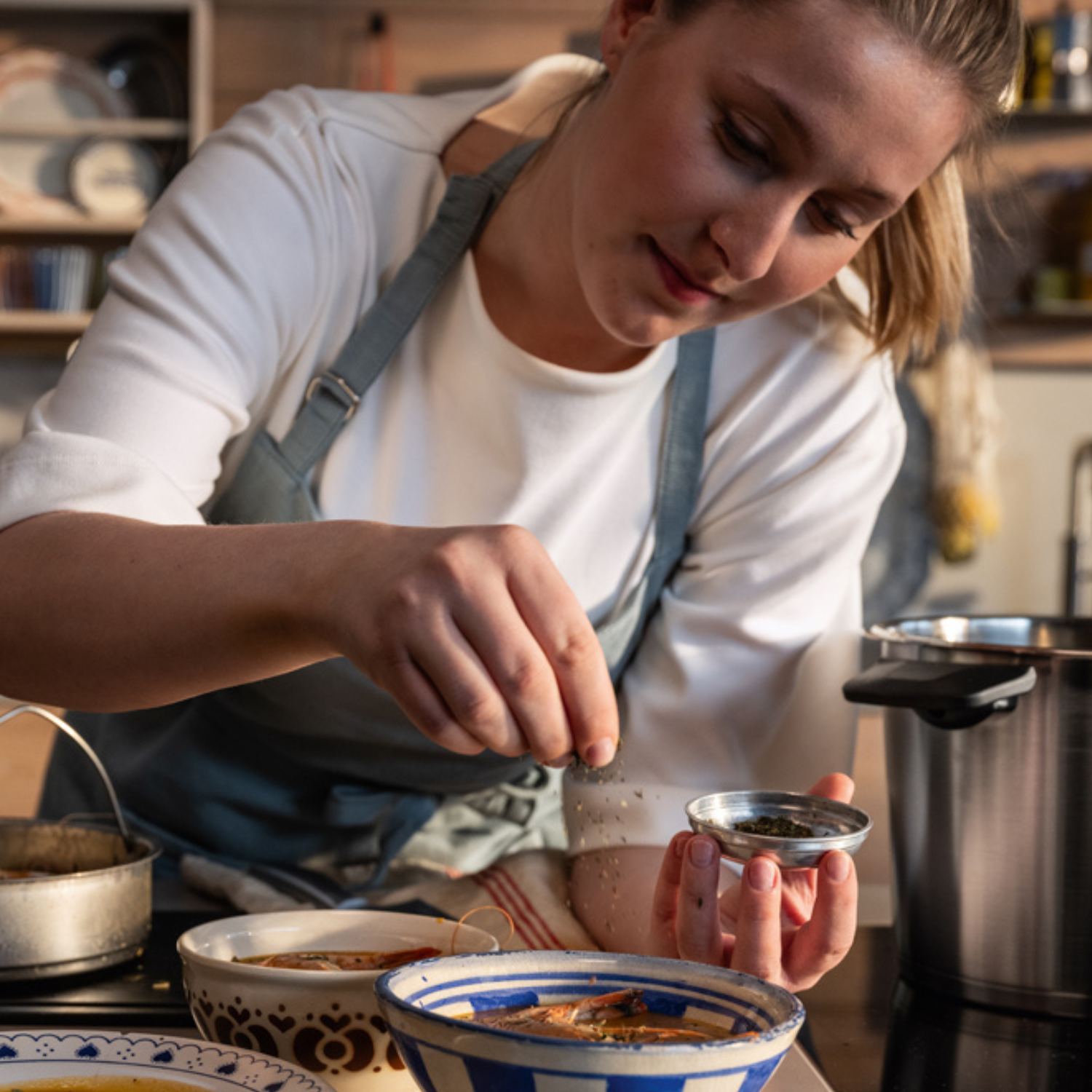 A person in an apron sprinkles seasoning over a bowl of food in a kitchen, surrounded by premium Fissler stainless steel pots and cookware. The scene reflects Fissler’s tradition of precision engineering, timeless design, and innovation “Made in Germany.”.