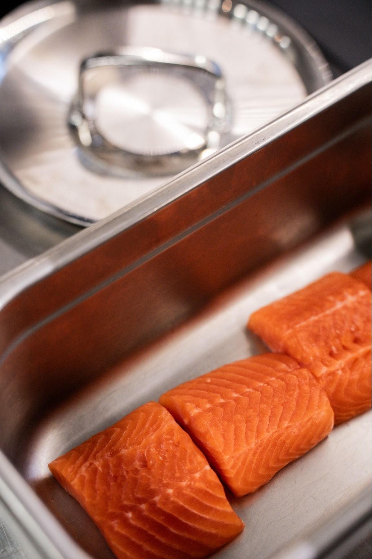 Three fresh salmon fillets lie in a Fissler stainless steel tray, behind which our iconic pot and lid, crafted with German precision, are hidden.