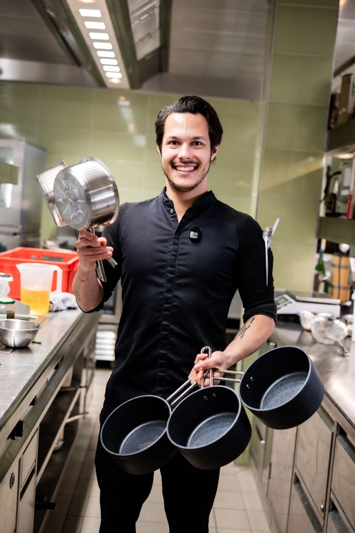 Smiling chef in black holding premium Fissler stainless steel cookware in a modern kitchen with green tiled walls.