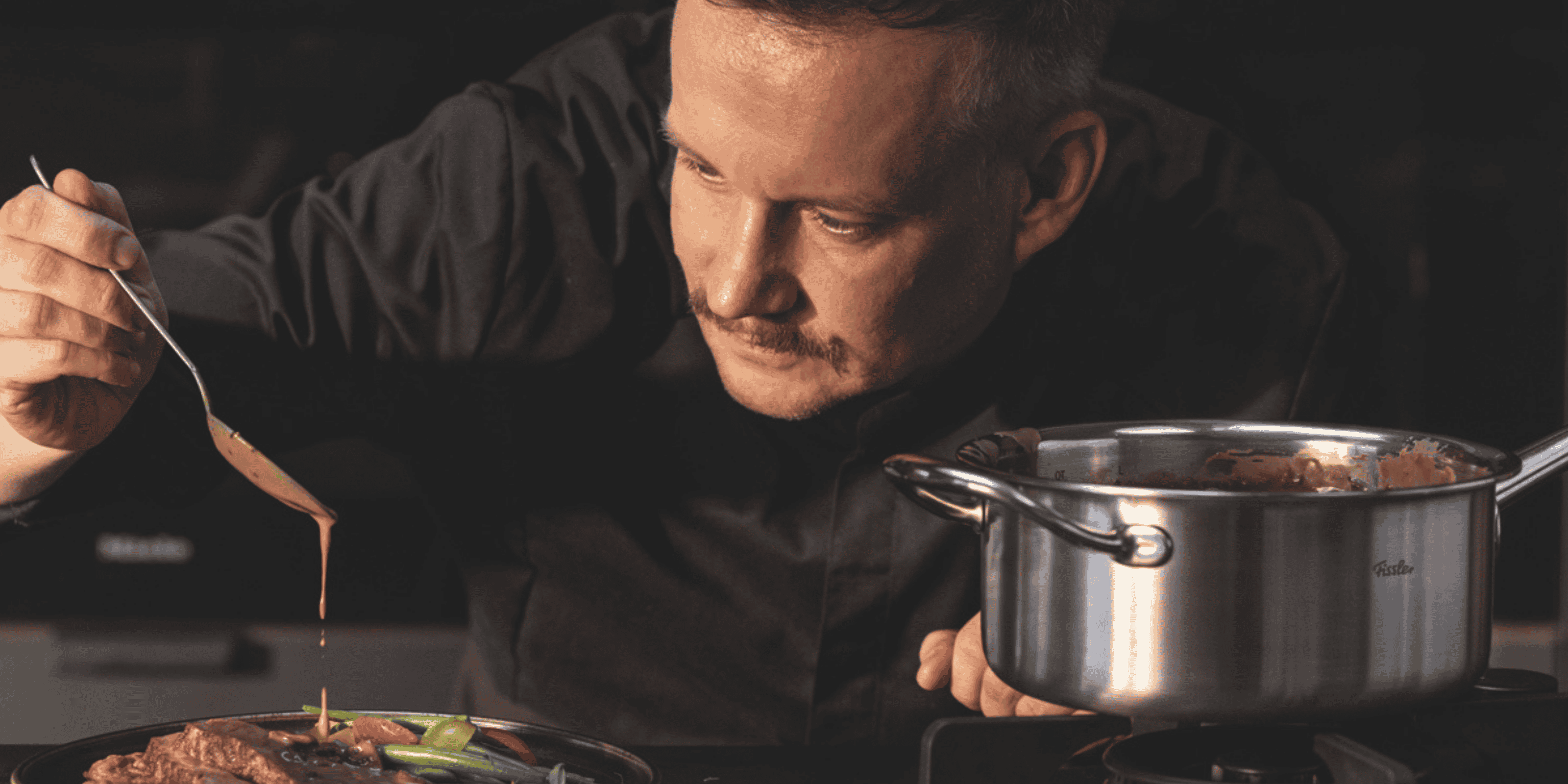 Ein Koch in dunkler Uniform gießt mit Präzision Sauce über ein Gericht, neben einem Fissler-Edelstahltopf auf dem Herd, konzentriert und aufmerksam.